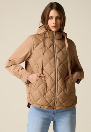 Kobieta ubrana w beżową pikowaną kurtkę zapinaną na zamek z kapturem i długimi rękawami, w połączeniu z jasnoniebieskimi dżinsami, stojąca na tle beżowego tła.