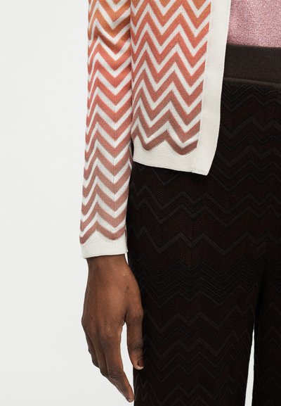 Κοντινό πλάνο ενός υψωμένου χεριού που φοράει τοπ με ροζ και λευκό ζιγκ-ζαγκ σχέδιο και σκουρόχρωμο παντελόνι με παρόμοια υφή ζιγκ-ζαγκ.