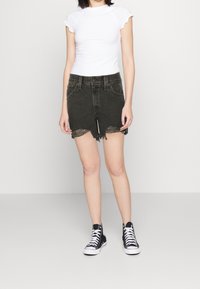 Top blanco de manga corta con bordes rizados, combinado con pantalones cortos de mezclilla verde oscuro con dobladillos deshilachados. Lleva puestas zapatillas altas negras.