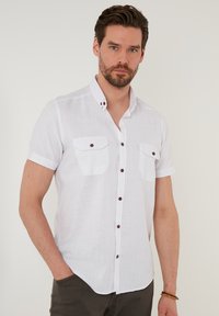 Camisa de lino blanca de manga corta con cuello abotonado, dos bolsillos en el pecho y botones marrones para contraste. Textura suave, corte relajado.