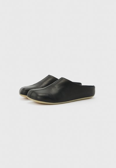 Ζευγάρι μαύρα δερμάτινα παντόφλες χωρίς κορδόνια με μπεζ σόλες, κλειστή μύτη και ανοιχτή πίσω πλευρά για εύκολη χρήση μέσα στο σπίτι.