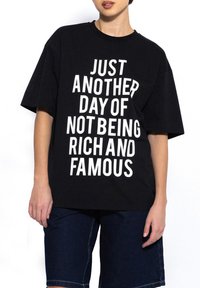 Czarny bawełniany t-shirt z wyraźnym białym napisem "Tylko kolejny dzień, w którym nie jestem bogaty i sławny", w luźnym kroju i krótkimi rękawami.