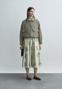 Woman wearing a cropped zip-up checked jacket over a cream midi skirt with green floral prints, holding a small black handbag, standing indoors.