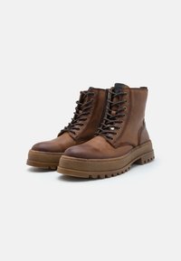 Botas de cuero marrón hasta el tobillo, con punta redondeada, diseño con cordones y suela de goma gruesa que presenta profundas ranuras para mayor tracción.