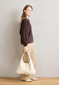 Brun langærmet bluse, beige bukser, holdende en hvid hobo taske. Blødt stof, løs design, enkle linjer og minimale accenter.