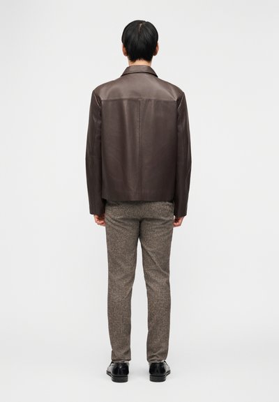 Άντρας που στέκεται πλάτη, φοράει σκούρο  καφέ δερμάτινο μπουφάν, γκρι υφασμάτινο παντελόνι και μαύρα επίσημα παπούτσια σε απλό λευκό φόντο.