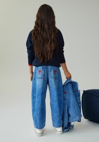 Personne aux cheveux bruns longs, vue de dos, portant un jean bleu avec une broderie de nœud rouge sur les poches, tenant une veste en denim assortie.