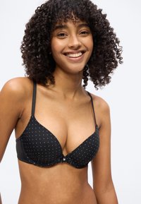 Νεαρή γυναίκα με σγουρά μαλλιά που χαμογελά, φοράει μαύρο σουτιέν με μικρές λευκές πουά σε απλό ανοιχτό φόντο.