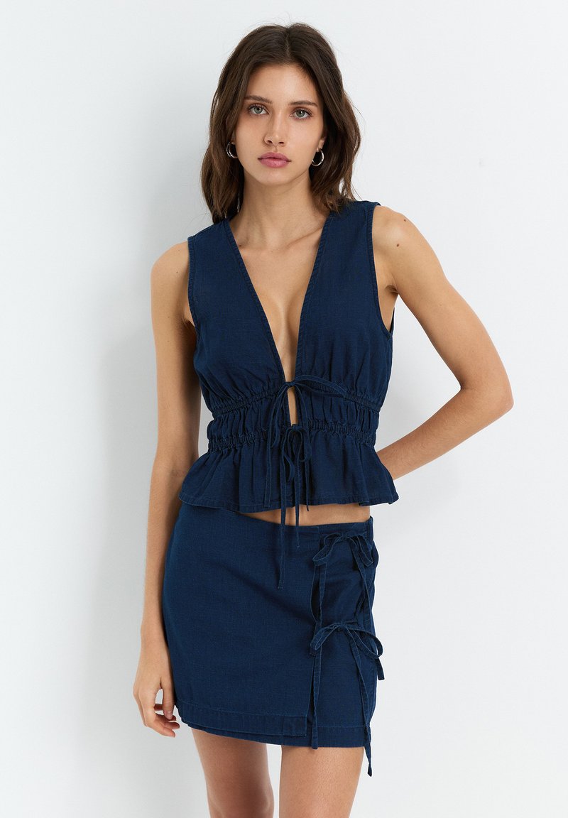 Haut sans manches bleu marine avec un décolleté en V profond, taille cintrée et ourlet volanté, assorti à une mini-jupe avec liens latéraux.
