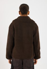 Bruine, gestructureerde fleecjas met een brede kraag, lange mouwen en een losse pasvorm. De achterkant laat schone naden en minimale details zien.
