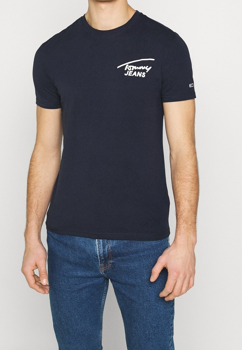 Uomo che indossa una t-shirt navy blue di Tommy Jeans e jeans blu, in piedi contro uno sfondo neutro, visibile solo il torso.