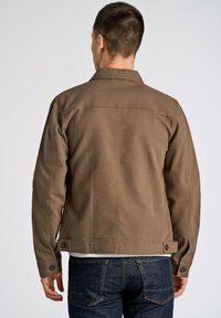 Brun canvasjacka med en figurnära design, knäppta ärmslut och sömmar bak. Bärs med mörka jeans i denim som visar en ren, minimalistisk stil.