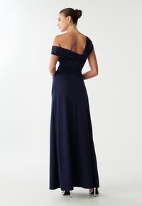 Granatowa sukienka wieczorowa z odkrytymi ramionami, z koralikowymi detalami, długości do podłogi i gładkiej struktury. Posiada ukryty zamek z tyłu.