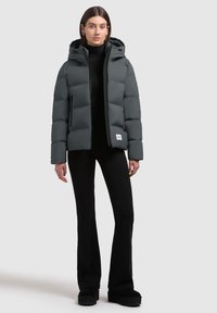 Chaqueta acolchada gris con capucha, que presenta un diseño acolchado, cremallera frontal y un pequeño parche de logo blanco en la parte inferior izquierda.