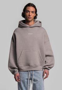 Felpa grigia oversize con tasca frontale e cordini, caratterizzata da un piccolo logo bianco sul petto. Realizzata in materiale morbido e testurizzato.
