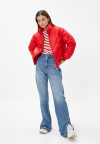 Rød pufferjakke med høj krave, parret med en stribet skjorte og brede blå jeans. Hvite sneakers og slidte kantdetaljer på jeansene.