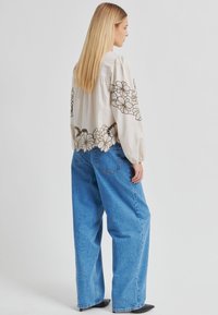 Blouse beige avec broderie florale marron, manches bouffantes, associée à un jean large en denim bleu et des chaussures à talons noires. Design simple, coupe décontractée.