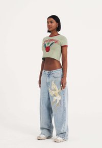 Lichtgroene cropped t-shirt met rode accenten en een kersenafbeelding. Losse blauwe jeans met een grote gouden vlinder borduursel op het been.