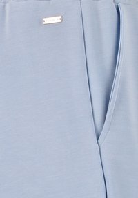 Hellbluse aus glattem Stoff, mit V-Ausschnitt und einem kleinen metallischen Logoakzent nahe der Schulter.
