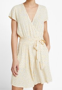 Robe jaune avec des manches courtes flottantes, décolleté en V profond et taille cintrée. Présente une ceinture à nouer et un motif à pois noir et blanc.