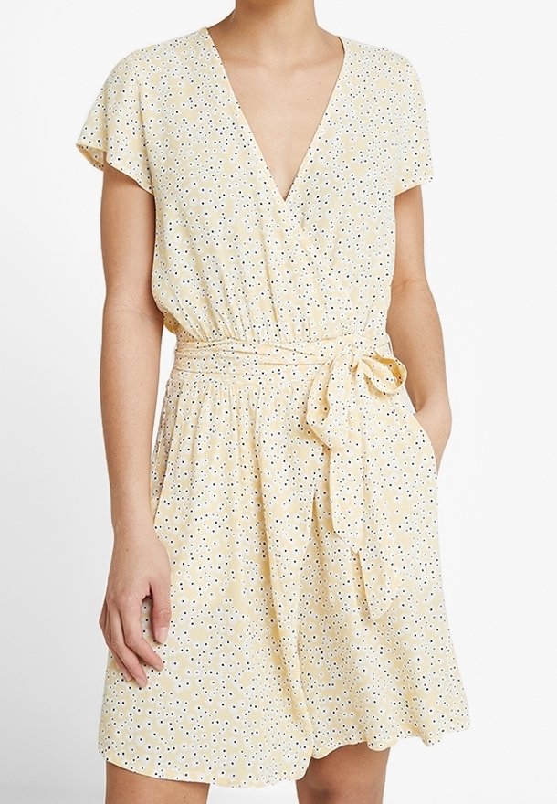 Robe jaune avec des manches courtes flottantes, décolleté en V profond et taille cintrée. Présente une ceinture à nouer et un motif à pois noir et blanc.