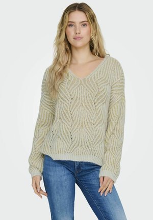 Hellgrüner Strickpullover mit V-Ausschnitt, der ein Wellenmuster und gerippte Bündchen aufweist. Getragen mit blauen Jeans; weiche Textur und lockere Passform.