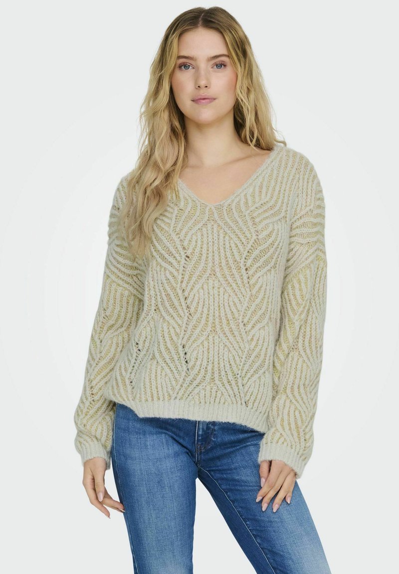 Hellgrüner Strickpullover mit V-Ausschnitt, der ein Wellenmuster und gerippte Bündchen aufweist. Getragen mit blauen Jeans; weiche Textur und lockere Passform.