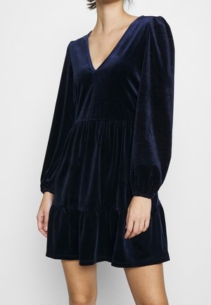 Granatowa sukienka z aksamitu z długimi bufiastymi rękawami, głębokim dekoltem w kształcie litery V i falbaną na dole. Miękka struktura i płynący fason podkreślają estetykę wizualną.