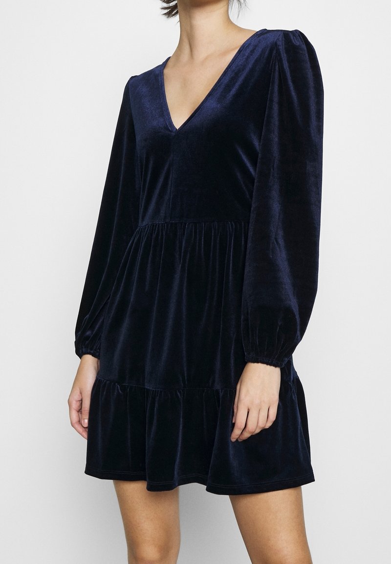 Robe en velours bleu marine avec manches longues bouffantes, décolleté en V profond et ourlet à volants. Texture douce et design fluide rehaussent l'attrait visuel.
