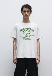 Hvid bomulds T-shirt med grøn grafisk tekst og billeder, herunder bjerge og en baseballspiller. Løs pasform med korte ærmer.
