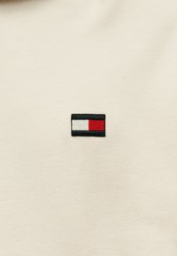 Ύφασμα κρεμ χρώματος με μικρό κεντητό λογότυπο που περιλαμβάνει ένα μαύρο ορθογώνιο, μια λευκή ρίγα και ένα κόκκινο τετράγωνο. Λείο υφή.