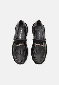 Paire de mocassins en cuir noir avec détails à franges et boucle argentée sur semelles crantées, vue de dessus sur fond blanc.