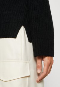 Schwarzer Strickpullover mit gerippter Textur und seitlichem Schlitz. Über einem cremefarbenen Stoff mit Taschenverschluss getragen.