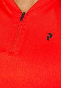 Gros plan d'un vêtement rouge zippé avec un logo noir sur la poitrine, montrant un tissu texturé et un col partiellement fermé.