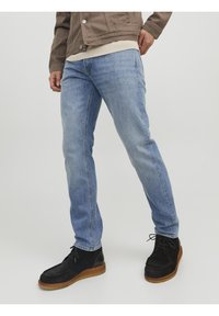 Jeans in denim azzurro chiaro con taglio dritto, indossati con stivali in suede neri con suola color cuoio. La giacca è marrone con un design a bottoni.