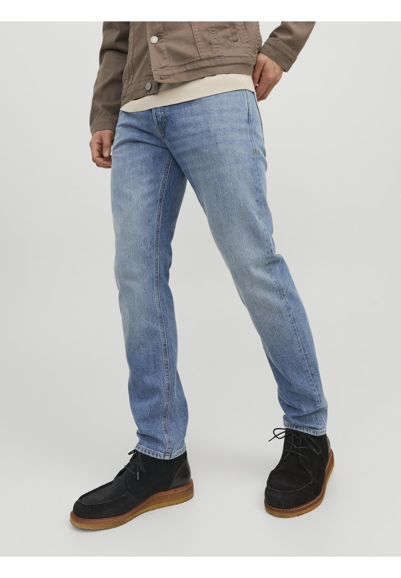 Jeans in denim azzurro chiaro con taglio dritto, indossati con stivali in suede neri con suola color cuoio. La giacca è marrone con un design a bottoni.