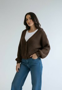 Brązowy dziergany kardigan z dekoltem w serek, guzikami z przodu i opuszczonymi ramionami, w połączeniu z jasnoniebieskimi jeansami z wysokim stanem, gładka faktura.