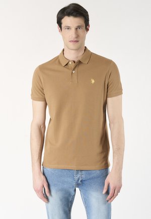 Uomo con polo marrone a maniche corte con piccolo logo giallo sul petto e jeans azzurri, in piedi contro uno sfondo uniforme.