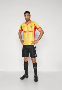 Vyras stovintis rankomis ant klubų, vilkintis geltonai raudonos spalvos dryžuotą futbolo marškinėlį, juodus šortus, juodas kojines ir futbolo batelius priešais vienspalvį foną.