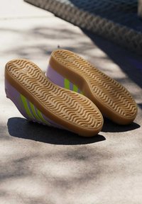 Par sneakers med lysebrune bølgede såler, lilla sider og gule striber, der ligger med sålerne opad på en solbelyst betonflade.