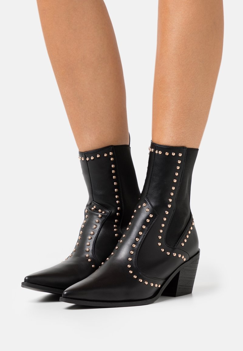 Bottines en cuir noir à bouts pointus, clous dorés le long des coutures et talons épais de hauteur moyenne, portées sur jambes nues.