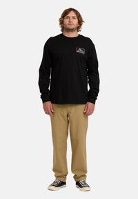 T-shirt nera a maniche lunghe con logo grafico frontale, abbinata a pantaloni beige chiaro e scarpe da skate nere con accenti bianchi.