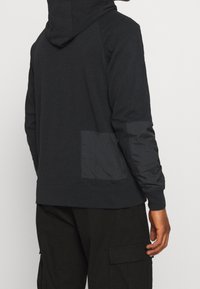 Mörk hoodie med en mjuk textur, en huva och en fyrkantig ficka med en annan tygtextur på nedre delen av ryggen.