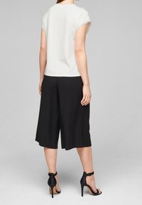 Bílý top s krátkým rukávem v kombinaci s černými culotami. Outfit má uvolněný střih a hladkou texturu. Vzhled doplňují černé sandály na vysokém podpatku.