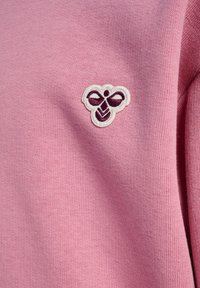 Roze trui met een zachte textuur, voorzien van een klein, wit geborduurd logo met een paarse accent aan de bovenlinkerkant.