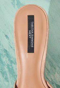 Semelle intérieure beige avec une étiquette rectangulaire noire affichant « Tabitha Simmons Next » sur une surface vert clair texturée.