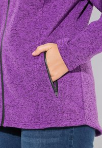 Lila fleece zip-jacka med texturerat tyg, sidofickor med dragkedja och en böjd nederkant. Materialet har ett fläckigt mönster.