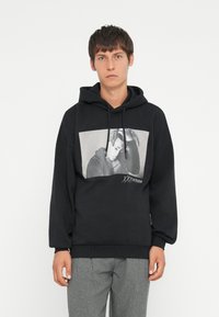 La felpa nera presenta una grande stampa grafica di una figura delineata in grigio. Include un cappuccio con cordino e polsini a costine, con una texture minimale.