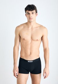 Schwarze Boxershorts mit einem weißen "BOSS"-Logo am elastischen Bund, getragen von einem männlichen Modell mit kurzen, dunklen Haaren vor einem hellen Hintergrund.