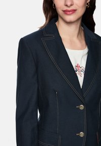 Navy blazer made of denim, featuring contrasting gold stitching, two buttons, and two front pockets. Inner shirt displays floral embroidery.
